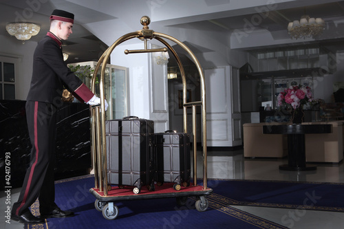 A uniformed doorman pushes a luggage cart .Several leather suitcases stand on luggage cart. Hotel service.The holiday season. Free space.