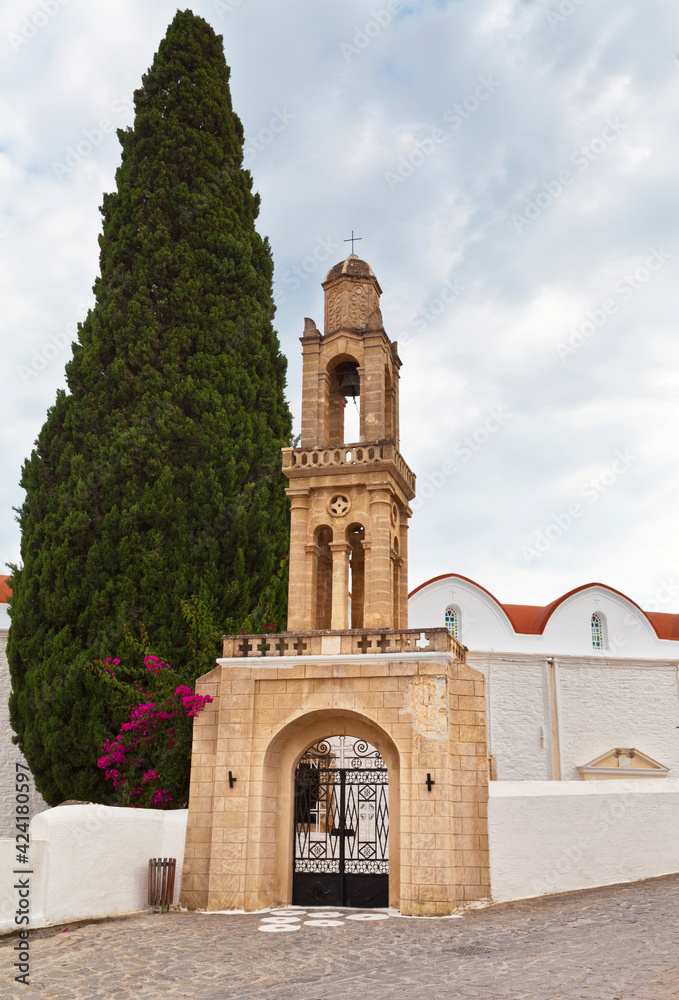Greece. Rhodes Island. Church of St. John the Evangelist in the village of Vati with a beautiful old stone bell tower and a cypress tree at the entrance on a summer day. Summer travel and excursions