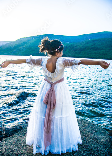 Una niña de espaldas y con los brazos en cruz contempla la inmensidad del Lago de Sanabria, España, vestida de Primera Comunión