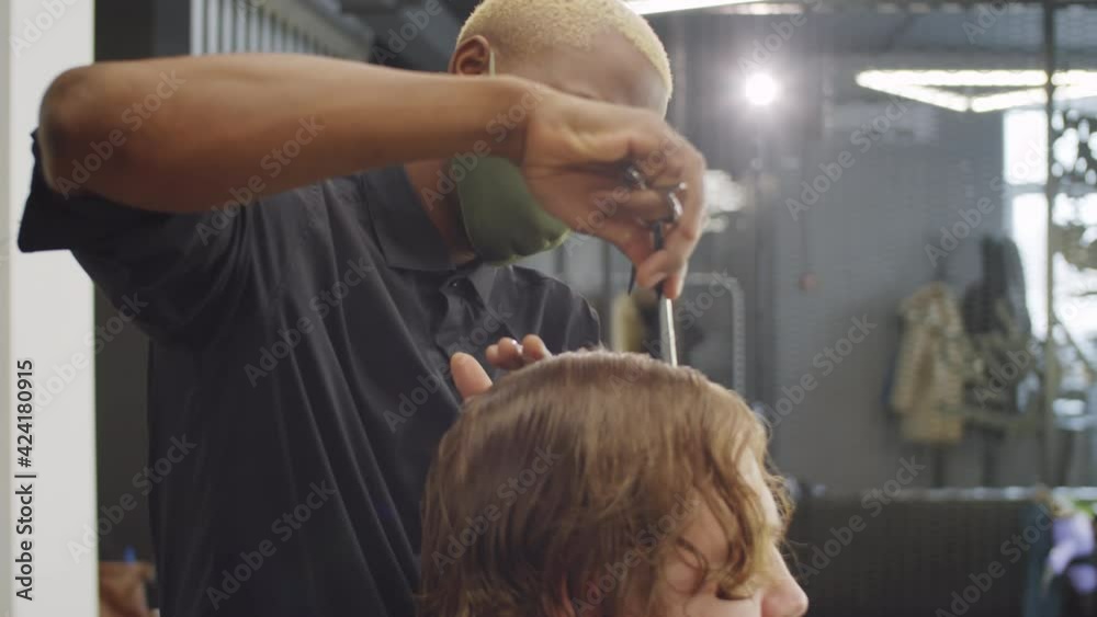 Afro-American barber in protective mask brushing wet hair of male client with comb before giving haircut while working in salon during coronavirus outbreak