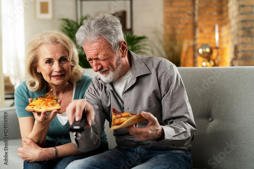  Cheerful husband and wife sitting on sofa at home. Happy senior woman and man eating pizza while watching a movie