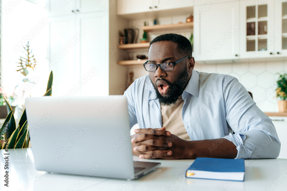 Fototapeta premium Shocked amazed African American man with glasses, freelancer, ceo or stock agent sits at his desk, looking at laptop in surprise, reading unexpected news or got big account profit