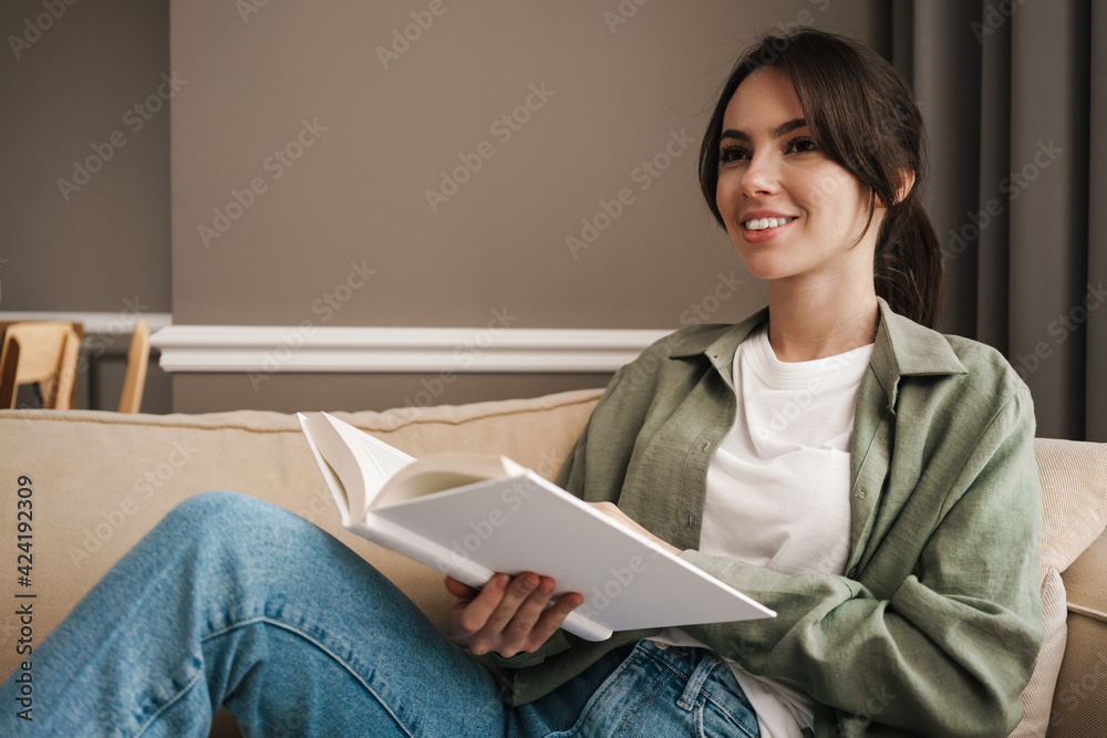 Obraz premium Portrait of a happy young woman sitting on couch with book