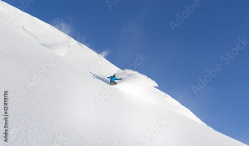 a cool snowboarder makes a turn with a plume of snow swirling on the edge of the blue sky and a large snow field. freeride high in the mountains-Heli skiing