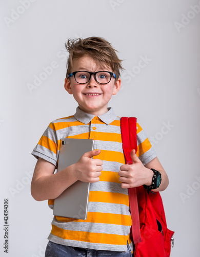 Portrait of happy school boy with backpack isolated against white background. Smiling face in glasses. Blond child in striped shirt with tablet in hands. Back to school