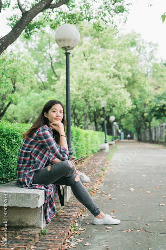 Smiling thoughtful young asian japanese woman crossed legs looking aside watching sits on bench in shade of tree in urban park. elegant lady with hands in chin waiting for someone outdoor in summer. photo