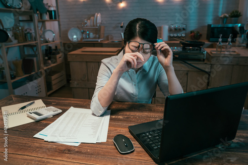 Feeling exhausted. Frustrated young asian japanese woman carrying eyeglasses and keeping eyes closed while sitting at working place at night time in home kitchen. female rubbing eyes by laptop pc.