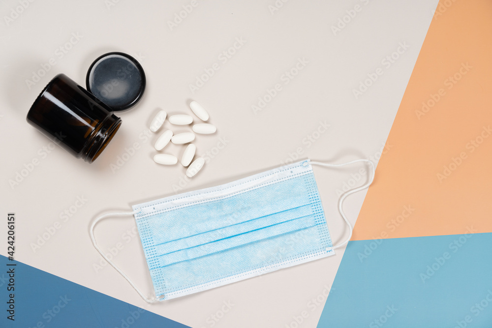 Glass jar with pills and medical mask on multicolored overlapped paper background. Concept, coronavirus, vitamins, health. Top view, copy space, close-up..fashionable still life, Glass jar with pills and medical mask on multicolored overlapped paper background. Concept, coronavirus, vitamins, health. Top view, copy space, close-up..fashionable still life,