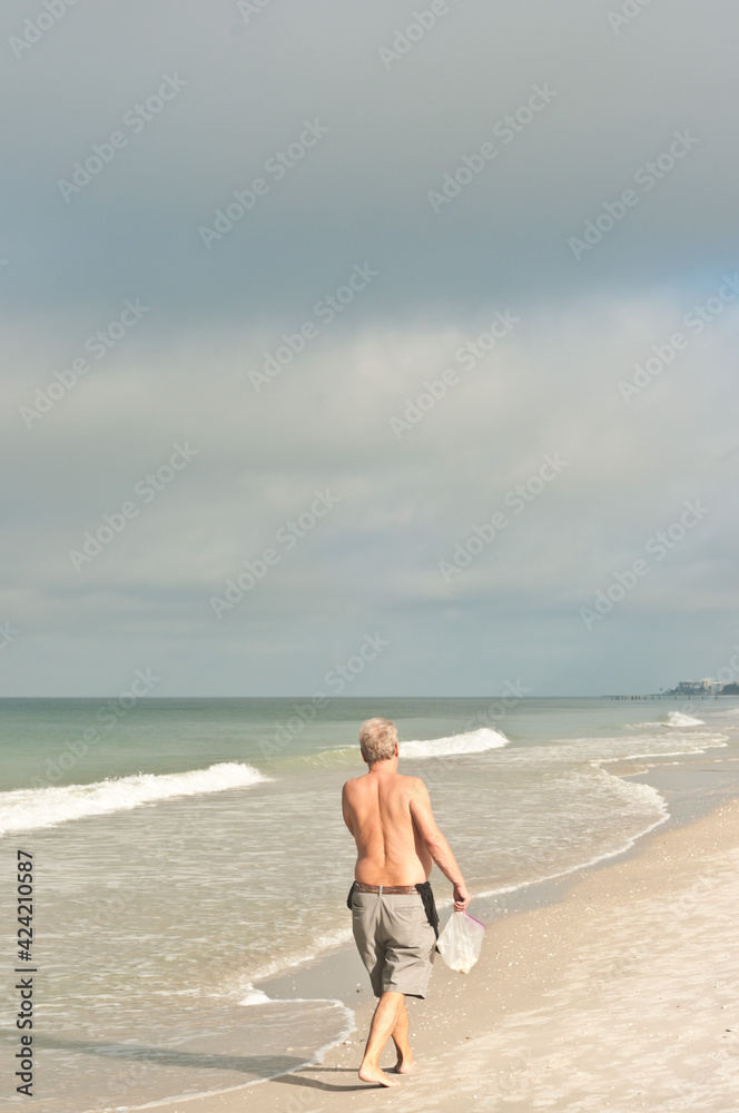 back view, far distance of a shirtless male walking along a sandy, tropical, shoreline, on an overcast day, with a streak of sunlight on him