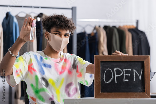 African american owner of showroom in protective mask holding hand sanitizer and chalkboard with open lettering