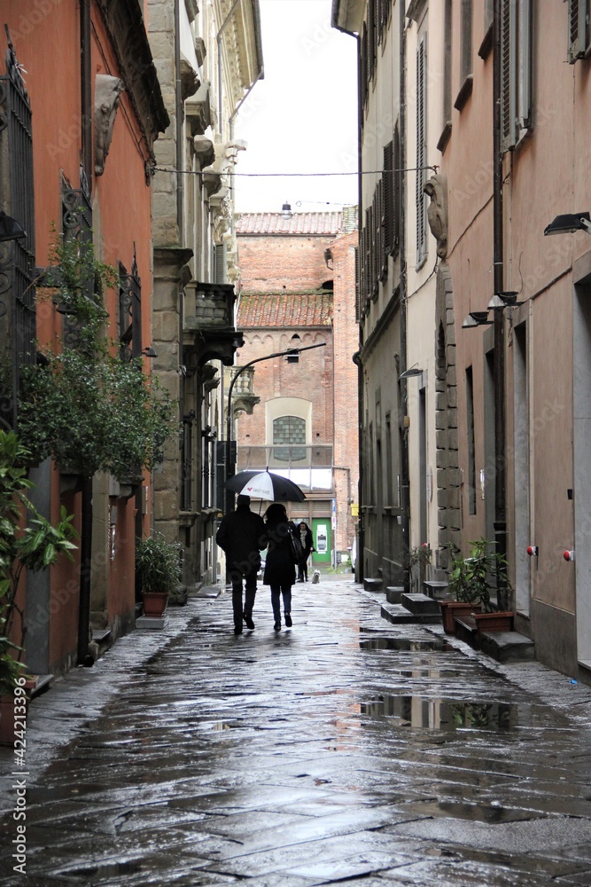 A well-dressed male female couple holding an umbrella walking down a wet lane, paved with large stone slabs, in the historic center of Pistoia, Italy, on a rainy early spring day