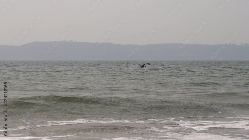 Pelican flying above the ocean waves flapping its wings in slow motion with coastal mountains in the background Pelican flying above the ocean waves flapping its wings in slow motion with coastal mountains in the background