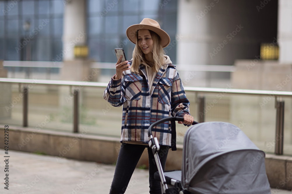 Happy Mother walking With Stroller In Park and using mobile phone. Joy of motherhood. Stylish young caucasian woman wearing warm clothes at autumn.