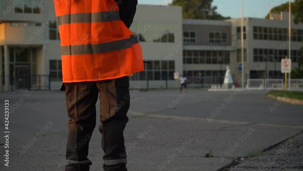 Cropped Image Of A Security Personnel In Orange Hi-Vis Watching A Kid Playing Football Outdoor. medium shot Cropped Image Of A Security Personnel In Orange Hi-Vis Watching A Kid Playing Football Outdoor. medium shot