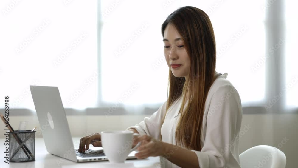 Young asian beautiful business woman working with laptop sitting at home drinking coffee. Smiling charming happy young female doing homework meeting conference with team at home. Young asian beautiful business woman working with laptop sitting at home drinking coffee. Smiling charming happy young female doing homework meeting conference with team at home.