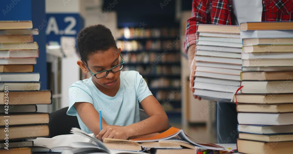 Librarian bringing stack of books to teen african schoolboy in library