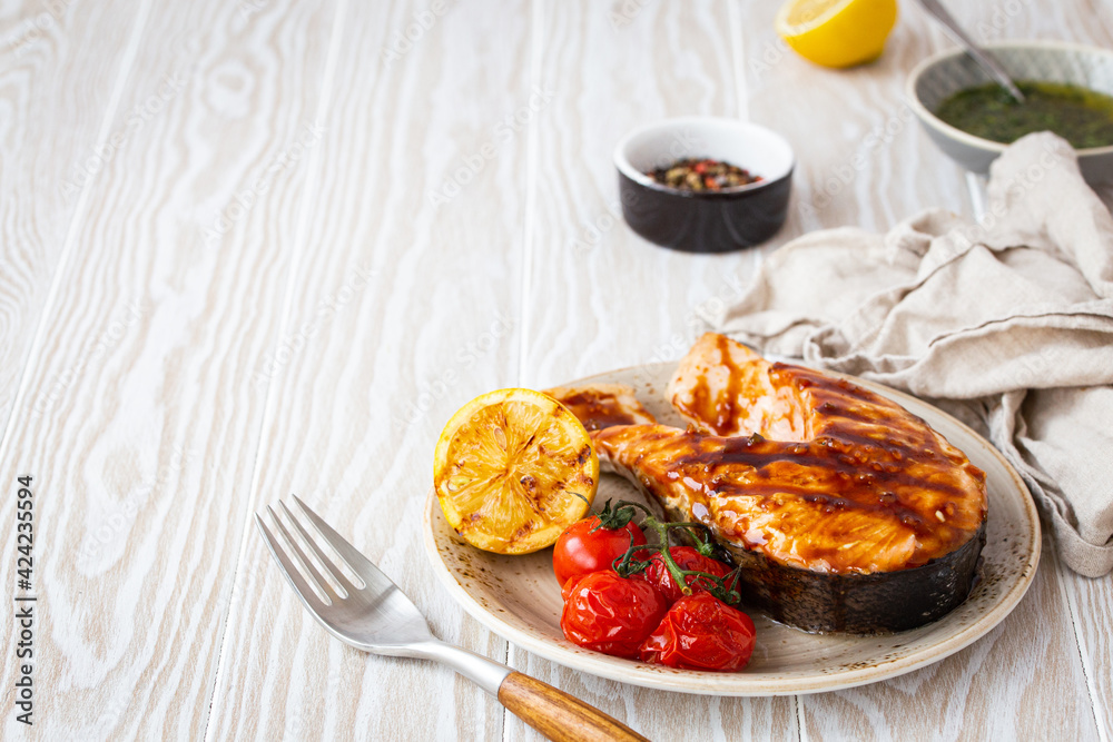Grilled salmon steak glazed with teriyaki sauce, vegetables and lemon served on ceramic plate on rustic white wooden table from angle view, selective focus with space for text