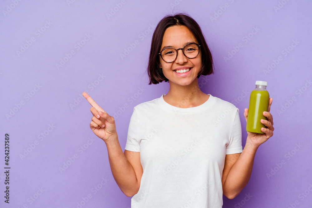 Young mixed race woman holding a healthy smoothie isolated on purple background smiling and pointing aside, showing something at blank space. Young mixed race woman holding a healthy smoothie isolated on purple background smiling and pointing aside, showing something at blank space.