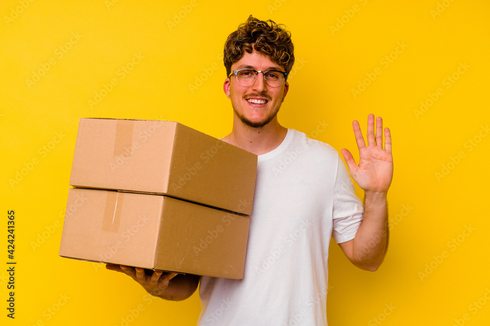 Young caucasian man holding a cardboard box isolated on yellow background smiling cheerful showing number five with fingers. Young caucasian man holding a cardboard box isolated on yellow background smiling cheerful showing number five with fingers.