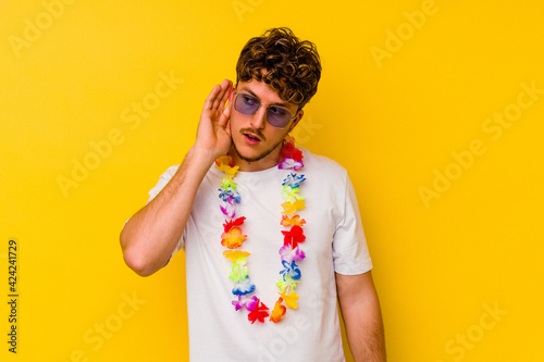 Young caucasian man wearing a Hawaiian party stuff isolated on yellow background trying to listening a gossip.
