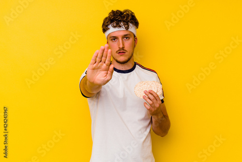 Young sport caucasian man eating a rice cake isolated on yellow background standing with outstretched hand showing stop sign, preventing you.
