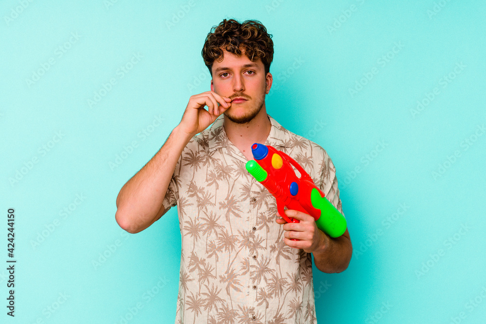 Young caucasian man holding a water gun isolated on blue background with fingers on lips keeping a secret.