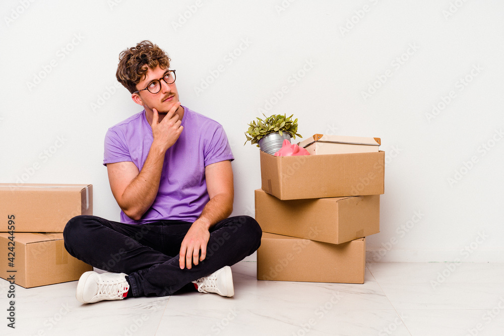 Young caucasian man sitting on the floor ready for moving isolated on white background looking sideways with doubtful and skeptical expression. Young caucasian man sitting on the floor ready for moving isolated on white background looking sideways with doubtful and skeptical expression.