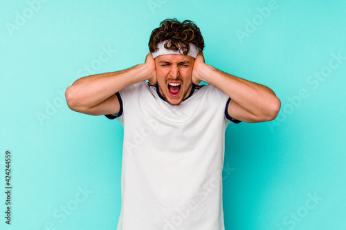 Young sport caucasian man isolated on blue background covering ears with hands trying not to hear too loud sound.