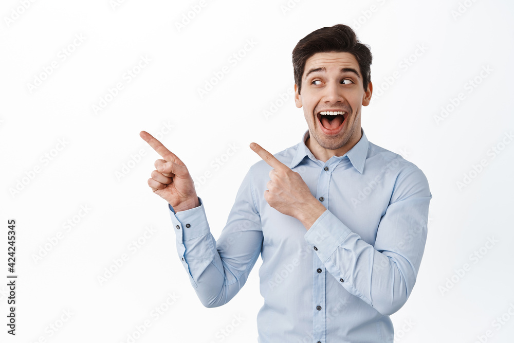 Cheerful businessman gasping fascinated, looking and pointing aside left, showing advertisement, standing over white background in blue collar shirt Cheerful businessman gasping fascinated, looking and pointing aside left, showing advertisement, standing over white background in blue collar shirt