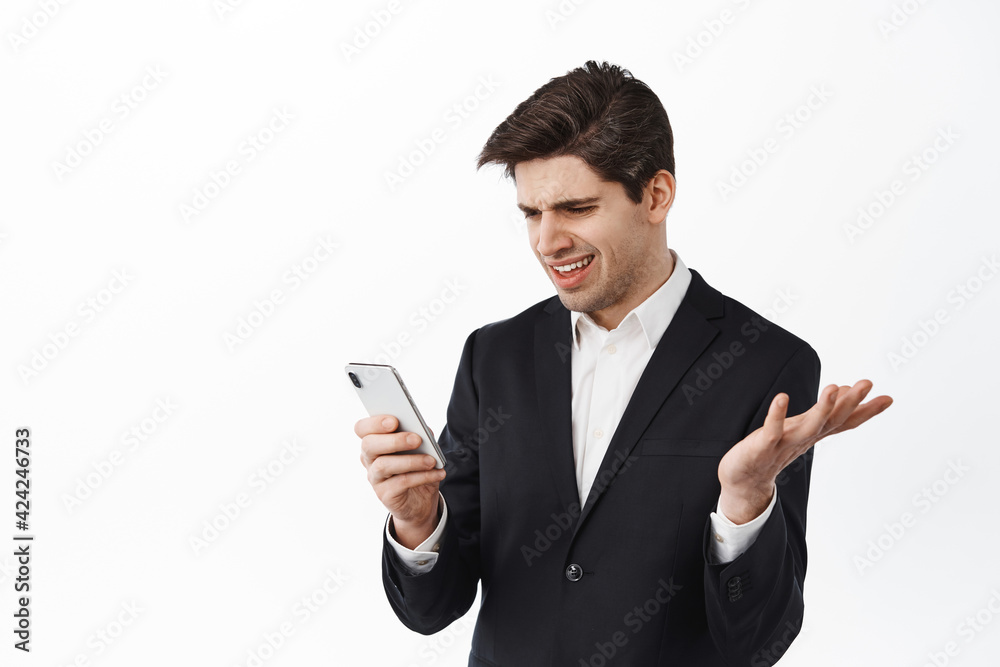Confused business man looking at smartphone with puzzled face, reading strange news message, standing against white background in black suit Confused business man looking at smartphone with puzzled face, reading strange news message, standing against white background in black suit