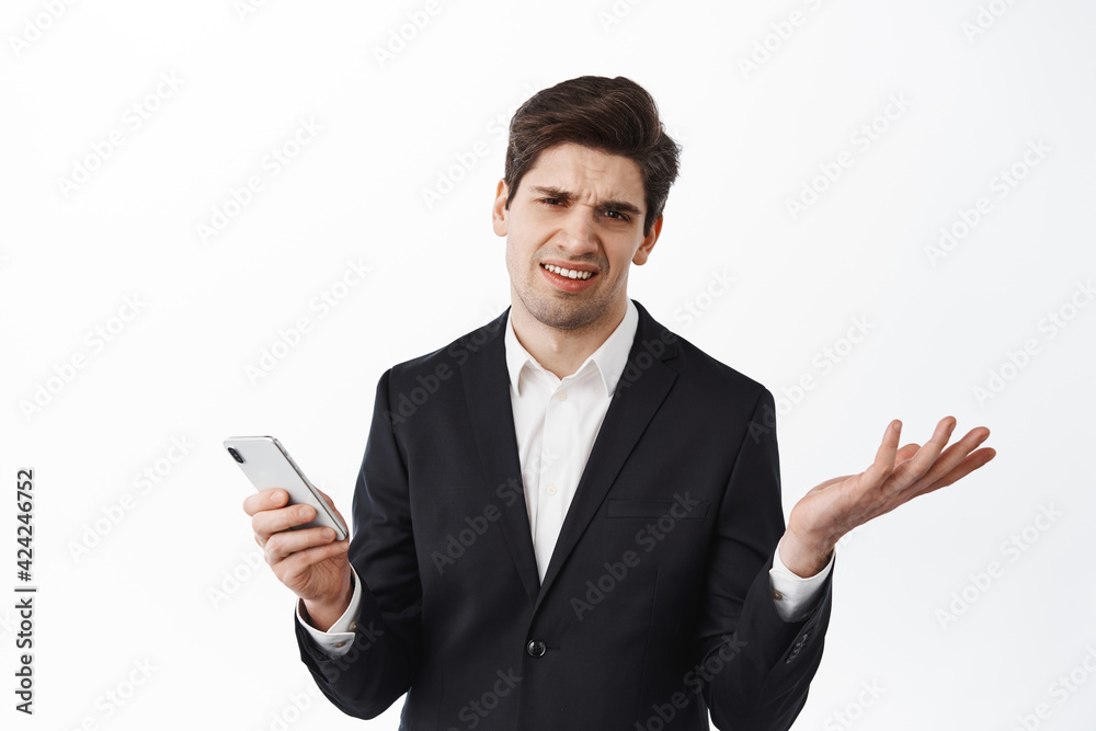 Bothered careless businessman shrugging shoulders and holding smartphone, dont care, cant understand something strange, standig over white background Bothered careless businessman shrugging shoulders and holding smartphone, dont care, cant understand something strange, standig over white background