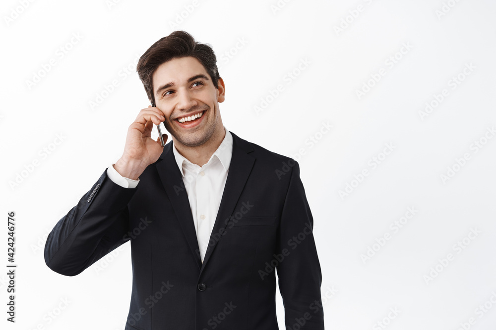 Handsome and confident businessman talking on mobile phone, smiling and looking at copy space, standing in black suit against white background Handsome and confident businessman talking on mobile phone, smiling and looking at copy space, standing in black suit against white background