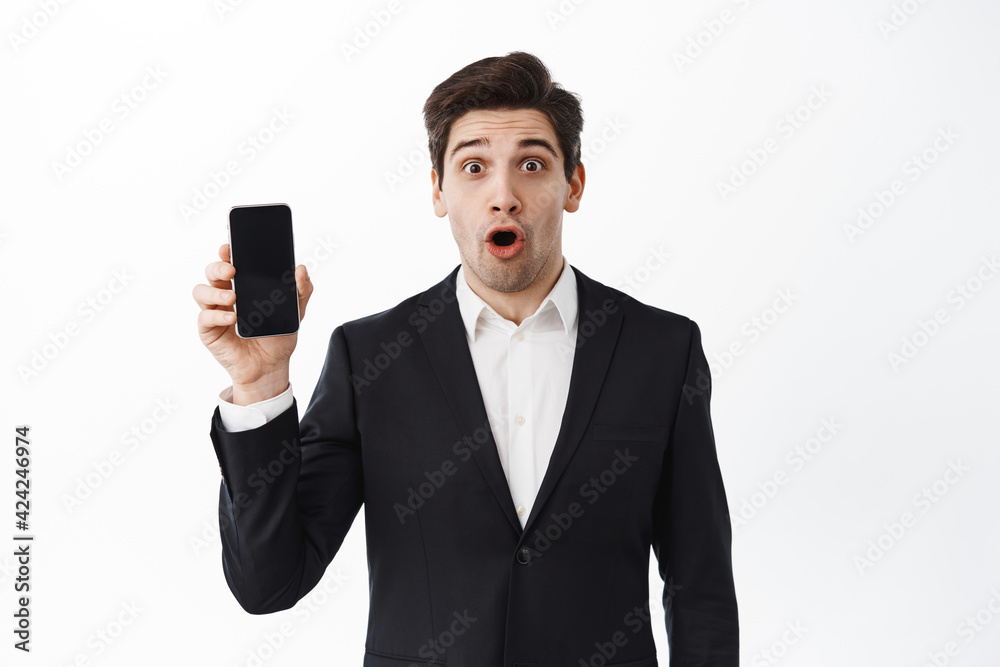 Surprised business manager in suit showing empty phone screen, make wow face and stare impressed, standing against white background Surprised business manager in suit showing empty phone screen, make wow face and stare impressed, standing against white background