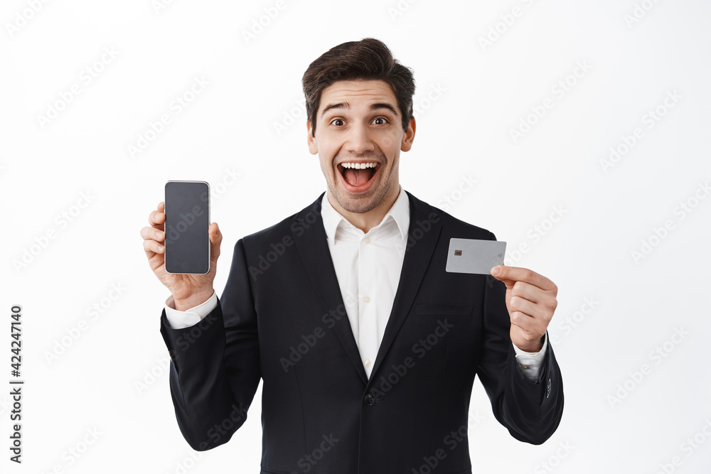 Surprised corporate man, businessman in suit shows empty phone screen and credit card, smiling amazed, new bank app, standing over white background Surprised corporate man, businessman in suit shows empty phone screen and credit card, smiling amazed, new bank app, standing over white background