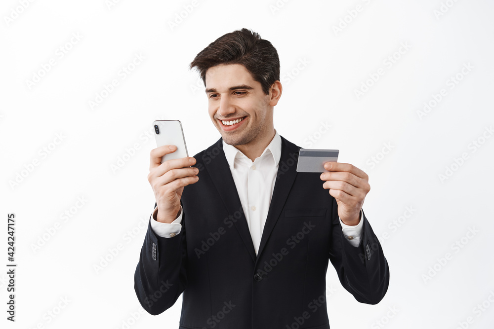 Smiling businessman paying online, show credit card and use mobile phone banking app, send money via application, shop online, standing over white background Smiling businessman paying online, show credit card and use mobile phone banking app, send money via application, shop online, standing over white background