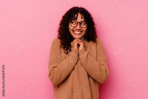 Young mixed race woman isolated on pink background keeps hands under chin, is looking happily aside. © Asier