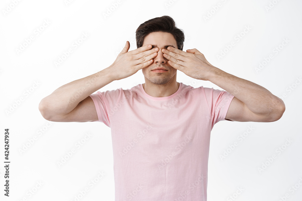 Serious man cover eyes with hands and waiting for something, anticipating surprise, standing against white background Serious man cover eyes with hands and waiting for something, anticipating surprise, standing against white background