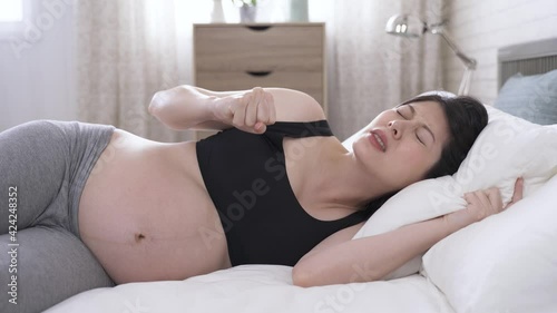 portrait japanese lady lying on bed is having labor contraction at home, and gripping pillow tightly and moaning with painful expression.