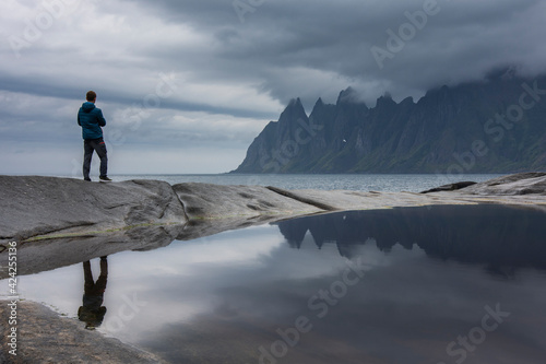 Tourist enjoys the magnificent view of Devil s Jaw or Devil s Teeth mountain chain  at coast at Tungeneset  Norwegian Sea. Gloomy weather  sky covered with grey clouds. Reflection like mirror. Senja.