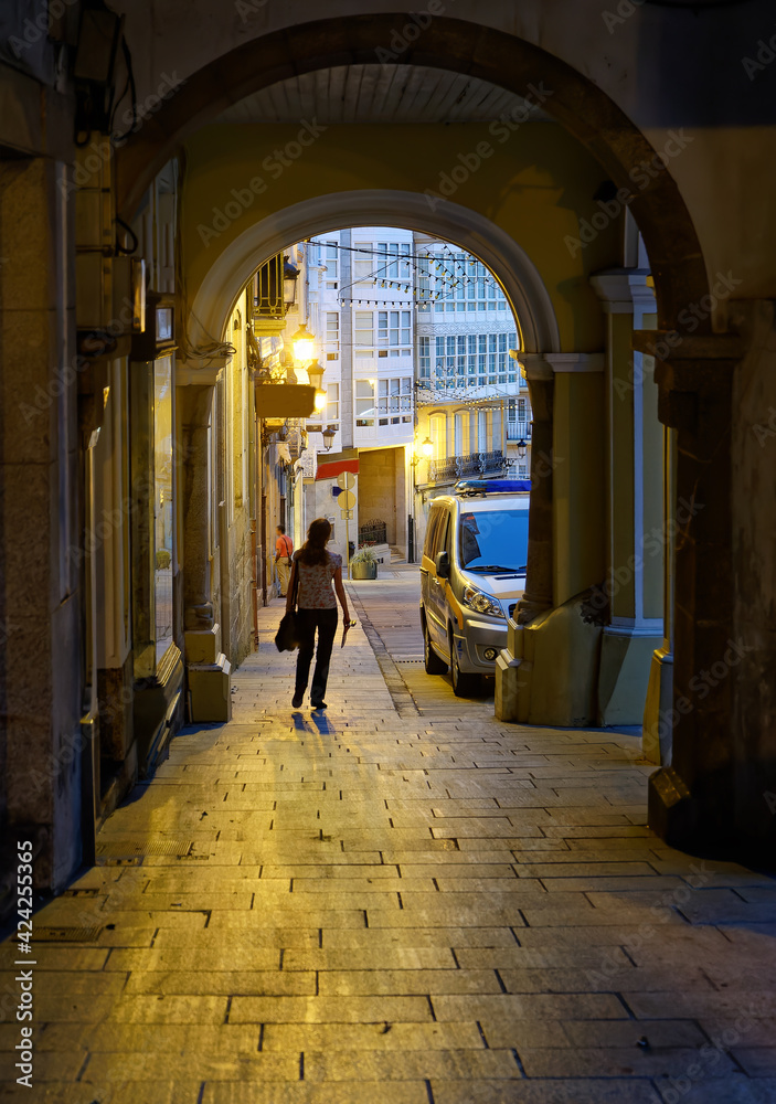 Silhouette of unrecognizable woman walking out of dark corridor in the historic town of Betanzos, Spain, with street lamps light reflecting on the floor stones.