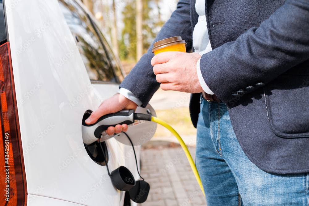 Close up man inserting power cable supply for charging electric car and holding cup of coffee. Close up man inserting power cable supply for charging electric car and holding cup of coffee.