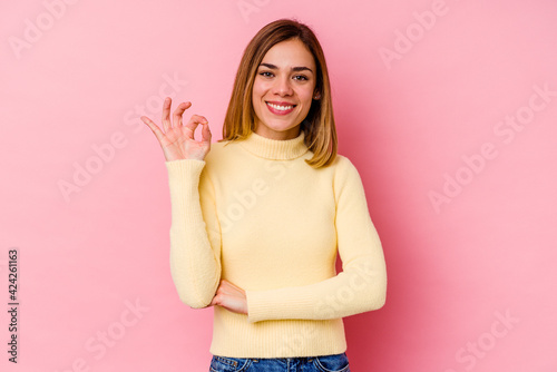 Young caucasian woman isolated on pink background winks an eye and holds an okay gesture with hand.
