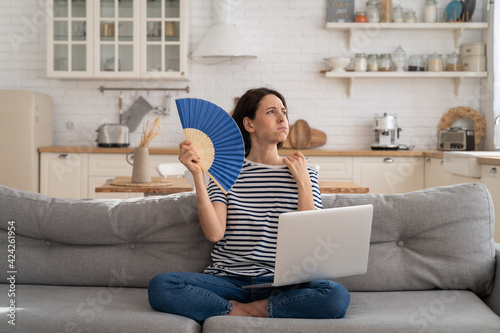 Tired millennial woman suffering from heatstroke flat without air-conditioner, waving blue fan, sitting on couch at home, working on laptop computer. Overheating, high temperature, hot summer weather.