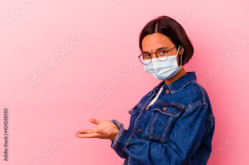 Young Indian woman wearing a mask antivirus isolated on pink background holding a copy space on a palm. © Asier