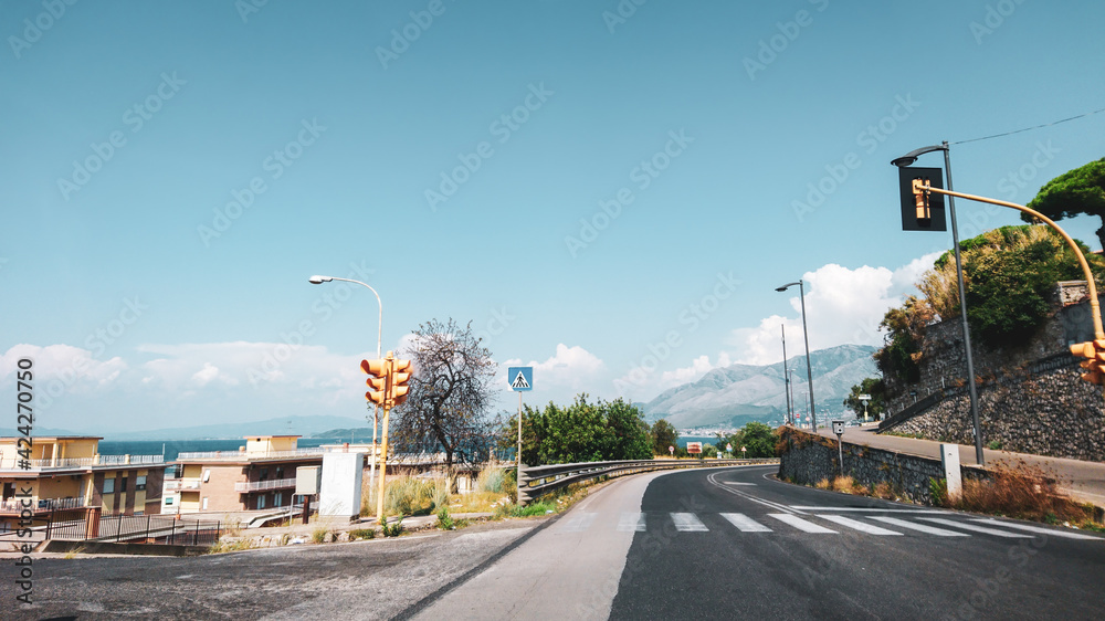 Driving empty road on Italian coast with crossroad, yellow traffic lights and blue clear sky. Scenic travel view in Mediterranean region. Color graded