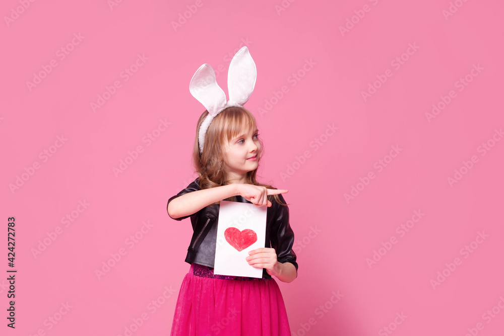 Cute blonde girl kid 7 y.o. with rabbit ears holding a card with red heart and points his finger to the side. Traditions, discounts, medicine, the concept of the celebration. Cute blonde girl kid 7 y.o. with rabbit ears holding a card with red heart and points his finger to the side. Traditions, discounts, medicine, the concept of the celebration.