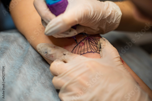 Master tattoo draws the red paint on the clients tattoo. Tattoo artist holding a pink tattoo machine in black sterile gloves and working on the professional blue mat. © MartaKlos