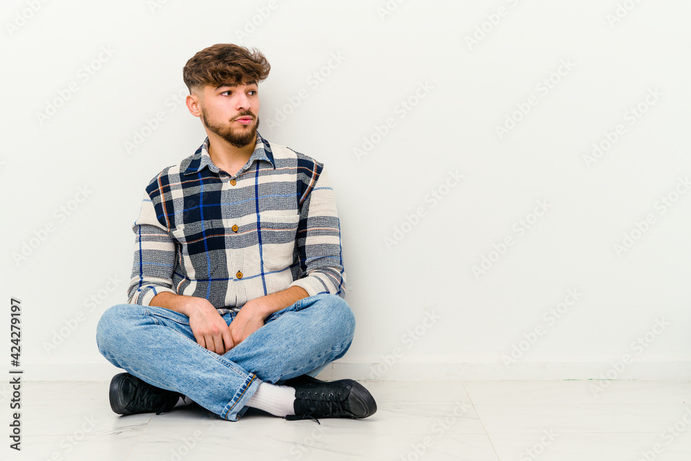 Young Moroccan man sitting on the floor isolated on white background looks aside smiling, cheerful and pleasant.