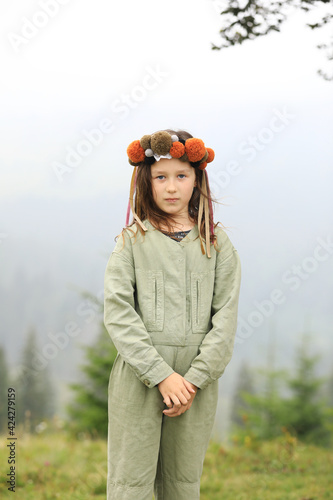 Cute little girl in a pastel jumpsuit with a colorful wreath on her head. Portrait of a girl in nature. A little girl outdoors in the foggy mountains looking in the camera. copy space © Andriy Medvediuk