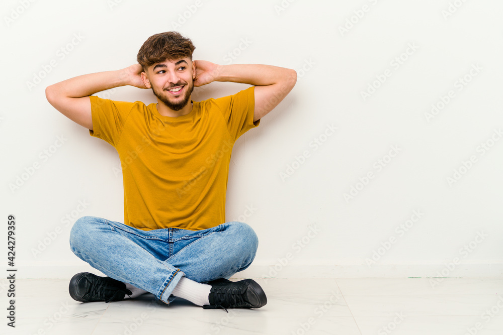 Young Moroccan man sitting on the floor isolated on white background feeling confident, with hands behind the head.